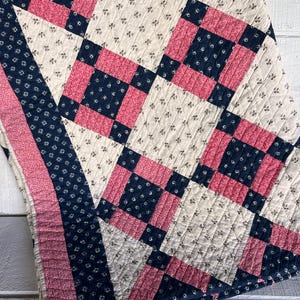 May include: A vintage quilt with a patchwork design. The quilt features a pattern of alternating squares in cream, navy blue, and pink. The navy blue squares have a small floral print. The quilt is folded, showing the detailed stitching.