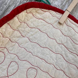 Peut inclure: Gros plan sur un tissu matelass&eacute; avec une bordure rouge et des coutures rouges ondul&eacute;es sur un fond blanc cass&eacute;. Une pince &agrave; linge en bois est attach&eacute;e au tissu. Le tissu fait probablement partie d'une courtepointe ou d'un textile plus grand.