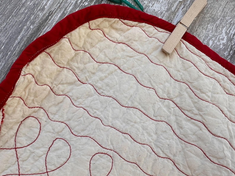 Peut inclure: Gros plan sur un tissu matelass&eacute; avec une bordure rouge et des coutures rouges ondul&eacute;es sur un fond blanc cass&eacute;. Une pince &agrave; linge en bois est attach&eacute;e au tissu. Le tissu fait probablement partie d'une courtepointe ou d'un textile plus grand.