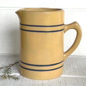 May include: A yellow ceramic pitcher with a smooth, glossy finish and a single handle. The cylindrical body features two sets of two horizontal dark blue stripes, one set near the top rim and one set near the base, with a pouring spout on the left.
