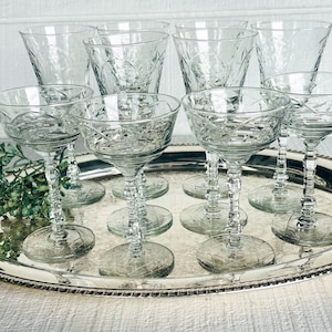May include: A set of eight clear crystal glasses, including champagne coupes and stemmed glasses, arranged on a silver serving tray. The glasses feature etched floral and geometric patterns. The tray is placed on a white surface.