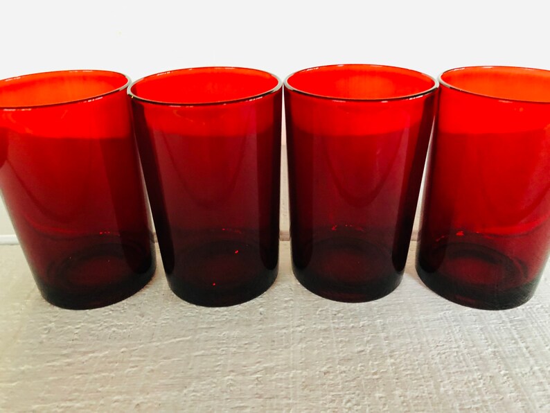 May include: Four red-tinted drinking glasses. The glasses have a cylindrical shape with a slightly tapered design. The rims of the glasses are a bright red, and the color fades to a darker red towards the base. The glasses are arranged in a row.