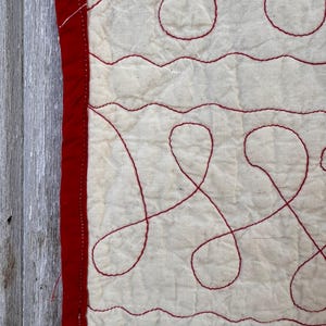 Peut inclure: Gros plan sur un textile matelass&eacute; avec un fond cr&egrave;me et des coutures rouges. Les coutures forment des lignes ondul&eacute;es et des motifs en boucle. Une bordure rouge longe le bord gauche du textile.