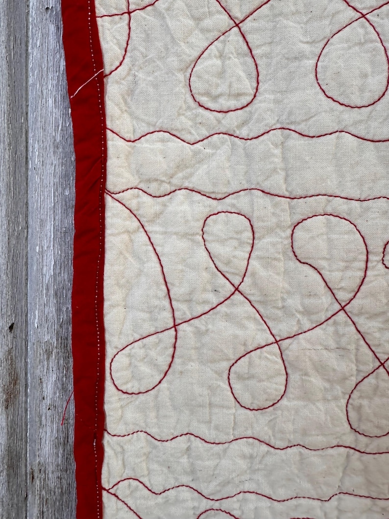 Peut inclure: Gros plan sur un textile matelass&eacute; avec un fond cr&egrave;me et des coutures rouges. Les coutures forment des lignes ondul&eacute;es et des motifs en boucle. Une bordure rouge longe le bord gauche du textile.