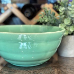 May include: A large, light green ceramic mixing bowl with a glossy finish. The bowl has a wide, rounded shape with several horizontal ridges. A small potted plant is in the background.