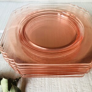 May include: A stack of pink glass dinner plates with scalloped edges. The plates are stacked on a white surface.