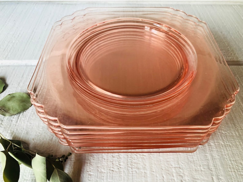 May include: A stack of pink glass dinner plates with scalloped edges. The plates are stacked on a white surface.