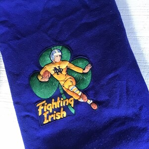 K&ouml;nnte beinhalten: Blaue Fleece-Decke mit einem gr&uuml;nen Kleeblatt und einem Cartoon-Footballspieler, der ein gelbes Trikot mit den Buchstaben "ND" tr&auml;gt. Der Text "Fighting Irish" befindet sich unter dem Kleeblatt.
