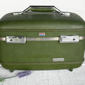 May include: A rectangular olive green hard-shell train case with a textured finish. It features a silver-tone metal band, two locking latches, and a top handle. The front displays a small American Tourister logo and a rectangular Tourister badge.