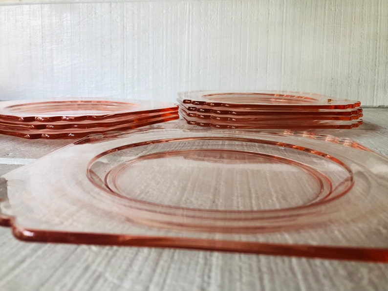 May include: A stack of six pink glass plates with scalloped edges. The plates are arranged in a pyramid shape, with the largest plate at the bottom and the smallest plate at the top.