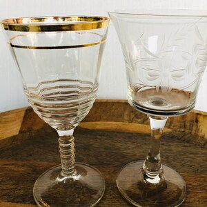 May include: Two clear glass wine glasses on a wooden surface. One glass has a gold rim and the other has an etched design.