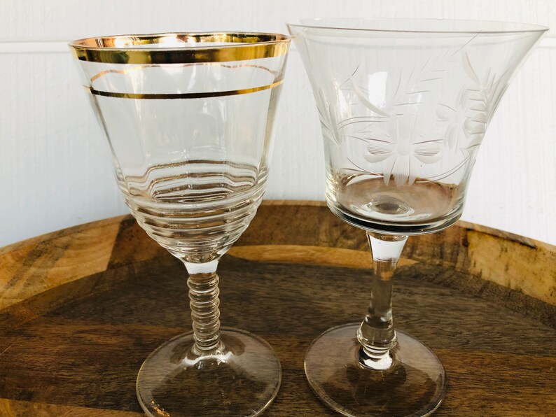 May include: Two clear glass wine glasses on a wooden surface. One glass has a gold rim and the other has an etched design.
