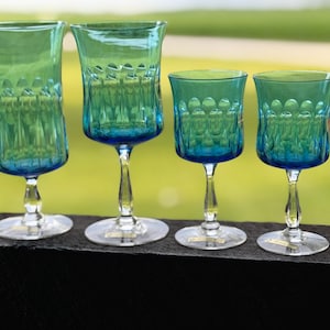 May include: A set of four teal blue glass goblets with clear stems and bases. Each glass features a distinct pattern of vertical indentations around the bowl, transitioning from a lighter teal at the top to a deeper blue hue at the base of the bowl.