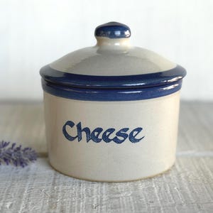 May include: A cylindrical ceramic cheese crock with a light beige body and a matching lid. The lid features a dark blue rim and a small knob handle. The word Cheese is printed in dark blue script on the front of the container.