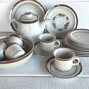 May include: A vintage white and brown ceramic dinnerware set. The set includes a teapot, serving bowl, cups, saucers, and plates. Some plates have a floral design. The rims and accents are brown.