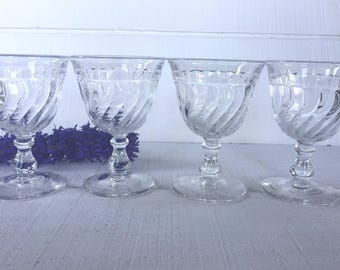 May include: Four clear, vintage-style stemmed glasses with a spiral design on the bowl. The glasses are arranged in a row, with a purple floral arrangement in the background. The glasses are made of glass.