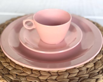 May include: A solid light pink ceramic tea set consisting of a cup with a circular handle, a small saucer, and a larger dinner plate. The pieces have a smooth, glossy finish and a simple, rounded design, stacked neatly in a vertical arrangement.