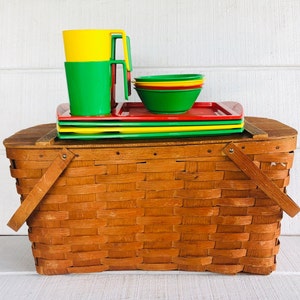 Puede incluir: Una cesta de picnic de mimbre marr&oacute;n con un juego de platos de pl&aacute;stico rojo, amarillo y verde y tazas a juego. El juego est&aacute; apilado encima de la cesta.