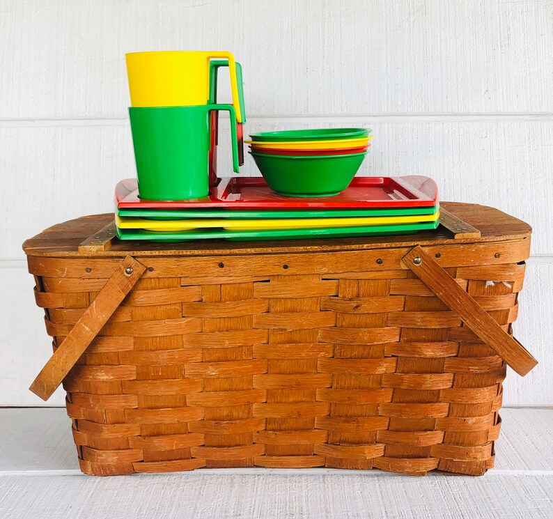 Puede incluir: Una cesta de picnic de mimbre marr&oacute;n con un juego de platos de pl&aacute;stico rojo, amarillo y verde y tazas a juego. El juego est&aacute; apilado encima de la cesta.