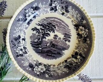 May include: A round, vintage plate with a cream-colored rim and a decorative edge. The plate features a central scene of a landscape with figures and a floral border in shades of gray and white. The plate is approximately 10 inches in diameter.