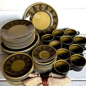 May include: A dinnerware set featuring olive green and dark brown ceramic plates, saucers, and mugs. The plates and saucers have a decorative border with a repeating geometric pattern. A tan fabric napkin with a wooden ring is in the foreground.