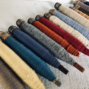 May include: A collection of spools of yarn in various colors, including beige, brown, blue, red, and orange. The spools are made of wood and have metal caps on the ends.