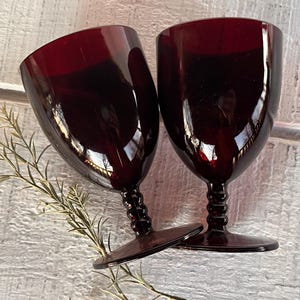 May include: Two ruby red wine glasses with a textured stem and a round base. The glasses are positioned on a white surface with a sprig of greenery. The glasses have a reflective surface.