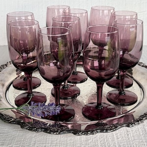May include: A collection of nine purple glass goblets with slender stems and rounded bowls, arranged on a round silver-toned metal tray with an ornate, scalloped edge. A small sprig of dried purple lavender rests on the tray surface.
