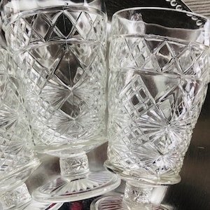 May include: Clear, vintage-style drinking glasses with a diamond pattern. The glasses are on a silver-colored tray. The glasses have a pedestal base and a wide rim. The diamond pattern catches the light, creating a sparkling effect.