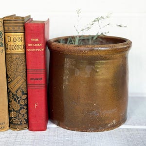 May include: A cylindrical brown ceramic crock with a glossy finish positioned next to three vintage hardcover books. The books are titled St. Elmo by Augusta J. Evans, Don Quixote, and The Golden Scorpion by Rohmer, featuring tan, gold, and red covers.