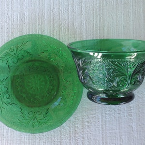 May include: A green glass bowl and saucer set. The bowl has a pedestal base and is decorated with floral and dot patterns. The saucer has a matching floral design. Both pieces are made of transparent green glass.