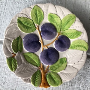 May include: A white ceramic decorative plate with a raised design of plums and leaves. The plums are purple, the leaves are green, and the branches are brown. The plate has a scalloped edge.