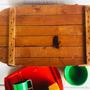Puede incluir: Una cesta de picnic de madera con tapa abatible y una mancha marr&oacute;n en la parte superior. La cesta est&aacute; abierta y se ven debajo vasos y bandejas de pl&aacute;stico rojo, verde y amarillo.