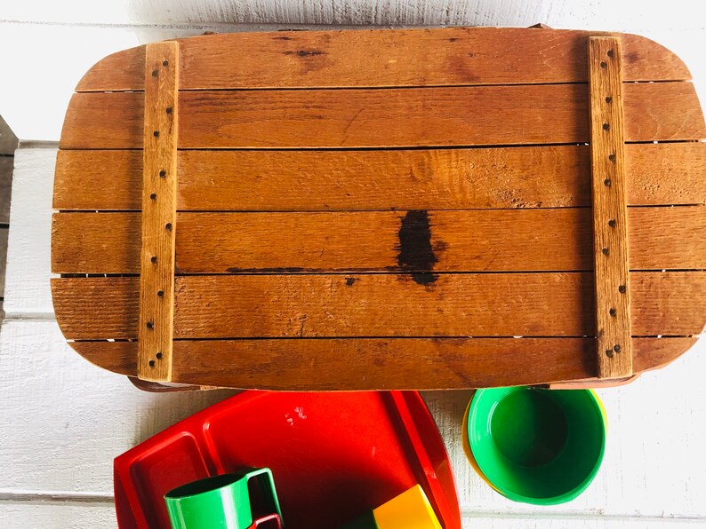 Puede incluir: Una cesta de picnic de madera con tapa abatible y una mancha marr&oacute;n en la parte superior. La cesta est&aacute; abierta y se ven debajo vasos y bandejas de pl&aacute;stico rojo, verde y amarillo.