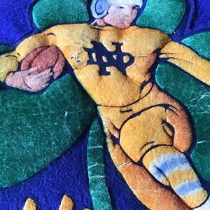 K&ouml;nnte beinhalten: Eine Vintage-Applikation aus blauem, gr&uuml;nem und gelbem Filz, die einen Footballspieler mit Helm zeigt, der einen Football h&auml;lt. Der Spieler tr&auml;gt ein gelbes Trikot mit den Buchstaben "ND" auf der Vorderseite. Der Spieler steht auf einem gr&uuml;nen Kleeblatt.