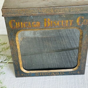 May include: Vintage metal biscuit tin with a rectangular window. The tin is painted in shades of blue and gold, with the words "CHICAGO BISCUIT CO" in gold lettering. The tin has a distressed, aged appearance.