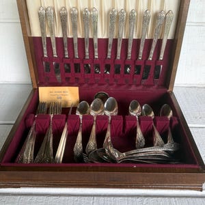 May include: A wooden storage chest with a cream and burgundy velvet interior containing a silver-plated flatware set. The set includes knives with ornate handles, forks, and spoons. A small yellow card reads 1847 ROGERS BROS. Original Rogers Silverplate.
