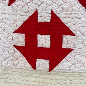 Peut inclure: Gros plan sur une courtepointe faite &agrave; la main avec un motif r&eacute;p&eacute;titif de formes rouges et blanches. La courtepointe pr&eacute;sente des motifs rouges en forme de fl&egrave;che et un fond blanc avec des coutures rouges en spirale. Le bord de la courtepointe est rouge.