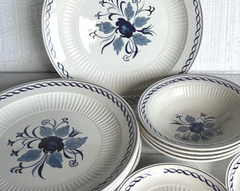 May include: A collection of white ceramic dinnerware with a blue floral design. The plates and bowls have a ribbed texture and a blue rope-like border. The floral design features a central blue flower with blue leaves.