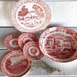 May include: A collection of antique red and white ceramic plates and bowls. The plates feature detailed landscape scenes and decorative borders. The set includes various sizes, perfect for serving a meal. A sprig of greenery adds a natural touch to the arrangement.