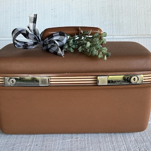 May include: A vintage brown suitcase with a handle, metal latches, and decorative stripes. A black and white checkered bow and greenery adorn the top. The suitcase is a classic travel accessory.
