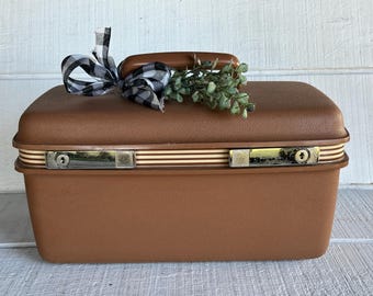 May include: A vintage brown suitcase with a handle, metal latches, and decorative stripes. A black and white checkered bow and greenery adorn the top. The suitcase is a classic travel accessory.