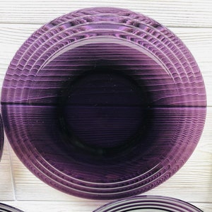 May include: A set of purple glass plates with a concentric circle design. The plates are stacked on a white wooden surface.