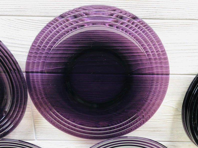 May include: A set of purple glass plates with a concentric circle design. The plates are stacked on a white wooden surface.