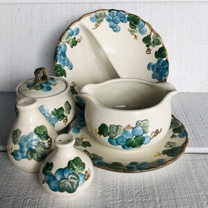 May include: A set of vintage ceramic serving dishes with a grapevine design. The set includes a gravy boat, a small plate, a sugar bowl, and two salt and pepper shakers. The dishes are decorated with blue and green grapes and leaves on a cream-colored background.
