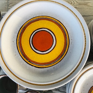 May include: A set of vintage ceramic dinnerware featuring plates and mugs. The plates have a light gray background with a central design of concentric circles in orange, yellow, and brown. The mugs are light gray with brown rims.