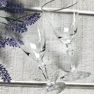 May include: Two clear, stemmed wine glasses with a delicate design. The glasses are angled, showcasing their elegant shape. Lavender sprigs add a touch of color against the white wooden background. Ideal for serving wine or other beverages.