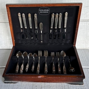 May include: A wooden silverware chest with a black velvet interior, holding a complete set of Holmes & Edwards Inlaid Silverplate flatware. The set includes knives, forks, spoons, and a serving spoon, all with ornate handles. The chest is open, revealing the neatly arranged silverware.