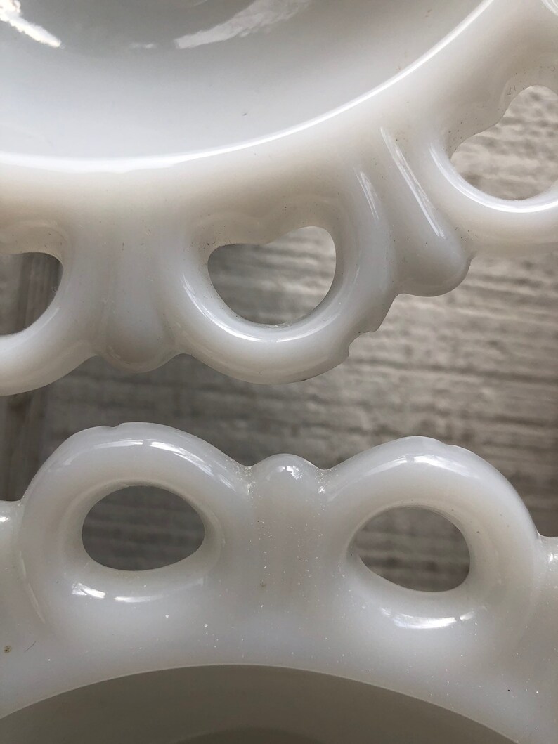 May include: White milk glass pedestal with a scalloped edge and openwork design. The pedestal has a smooth, glossy finish.