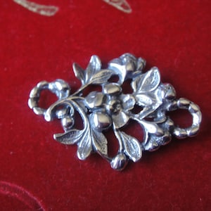 Puede incluir: Encanto de plata con un diseño floral detallado con hojas y bayas. El encanto tiene un bucle para sujetarlo a una pulsera o collar.